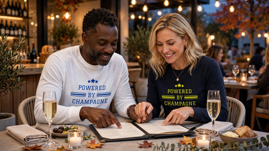 Happy couple at outdoor restaurant in autumn wearing matching Powered by Champagne long sleeve tees - white and navy Airlume combed cotton tees - pointing at menu together holding hands - champagne lovers gift idea for couples and best friends