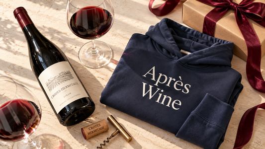 Folded navy blue Après Wine hoodie with white lettering, styled beside a bottle of Crozes-Hermitage Syrah red wine, two wine glasses, a corkscrew and a ribbon-wrapped gift box. A holiday wine and hoodie gift pairing from Gourmandee Tees.