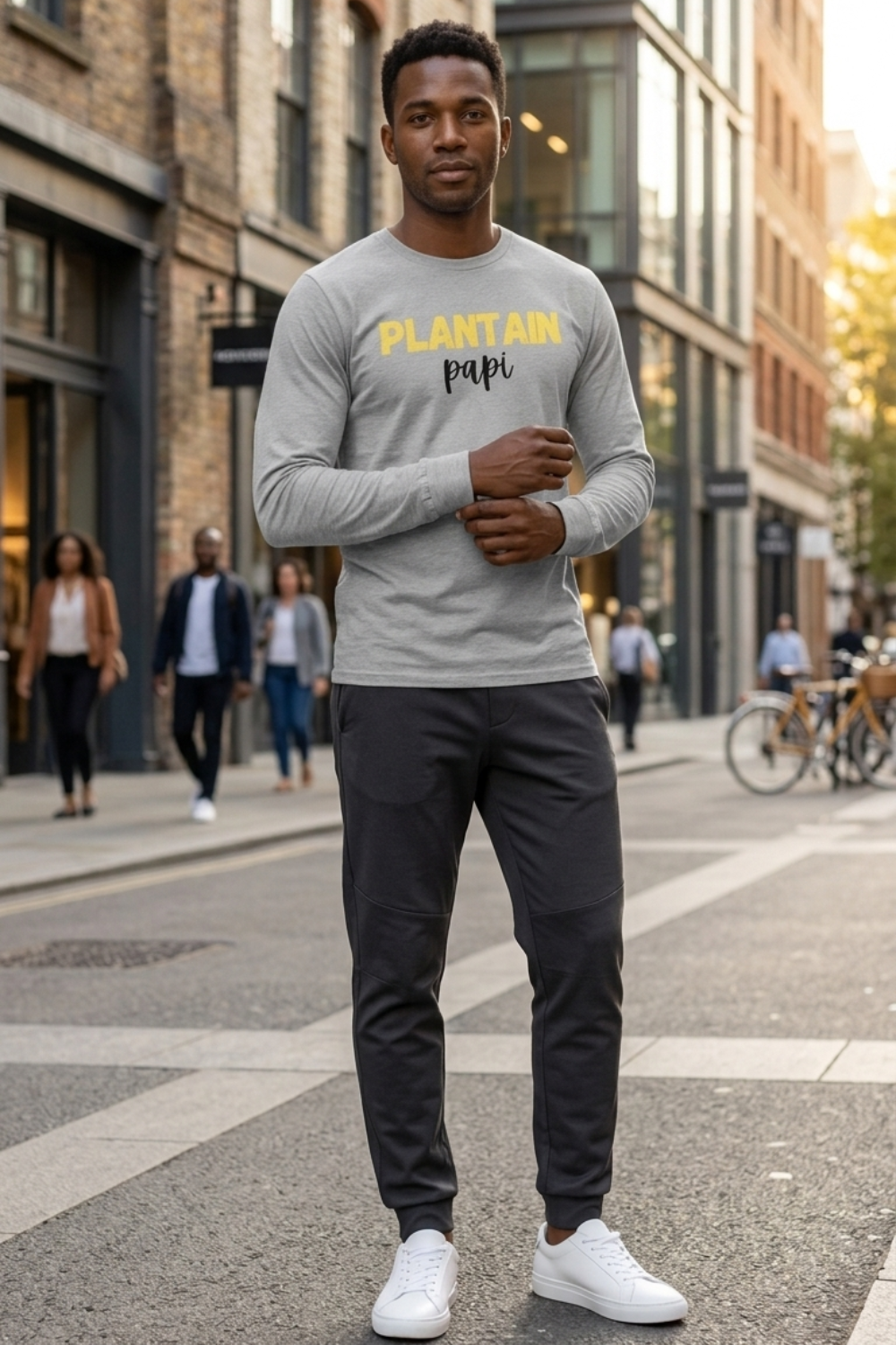 Black man wearing a heather grey Plantain Papi long-sleeve shirt with yellow and black lettering, styled with dark tapered track pants and white sneakers on a city street.