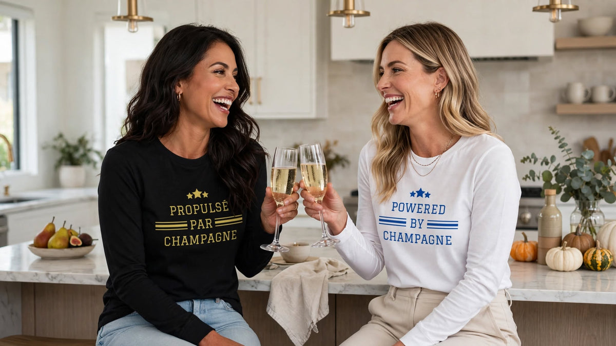 Two women laughing and toasting champagne in modern kitchen wearing Franglais champagne long sleeve gift set - black Propulsé Par Champagne tee and white Powered by Champagne tee - unisex Airlume combed and ring-spun cotton long sleeve tees for champagne lovers