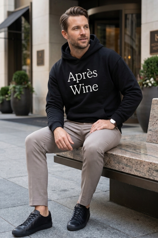 Man wearing a black Après Wine oversized wine lover hoodie with white serif lettering, styled with taupe trousers and black sneakers outside a modern hotel.