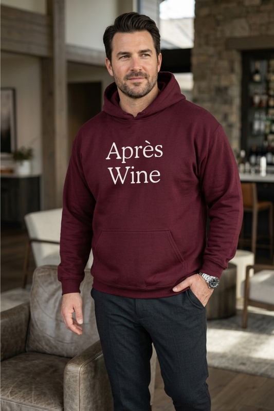 White male model wearing a maroon hoodie with Après Wine printed in white serif lettering on the front, styled with dark trousers in a modern hotel lounge.