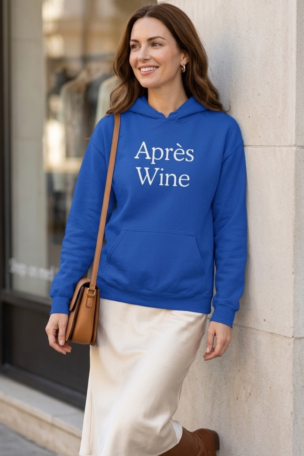 White woman wearing a royal blue Après Wine hoodie with white serif lettering, styled with a cream satin midi skirt and tan shoulder bag.