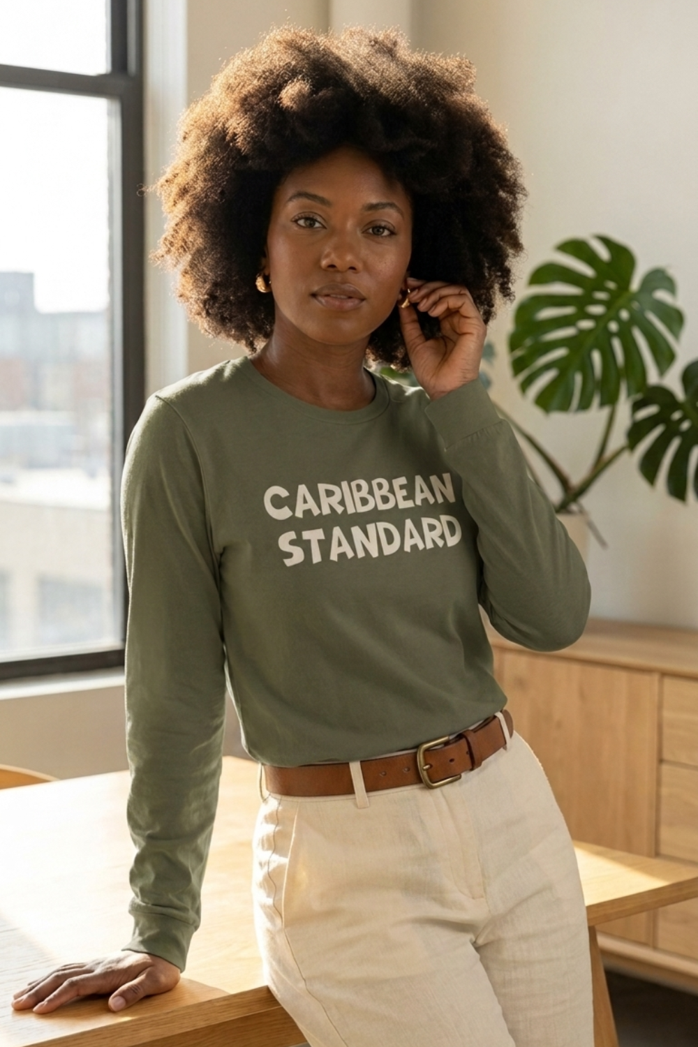 A woman with an afro hairstyle wearing an olive green long-sleeve t-shirt with 'CARIBBEAN STANDARD' printed in white across the chest, paired with white pants and a brown belt, standing in a bright room.