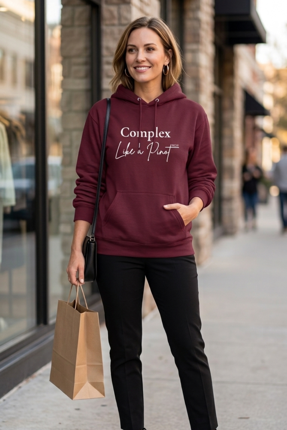 White woman wearing a burgundy Complex Like a Pinot Noir hoodie with white lettering, styled with black trousers and a crossbody bag while shopping.