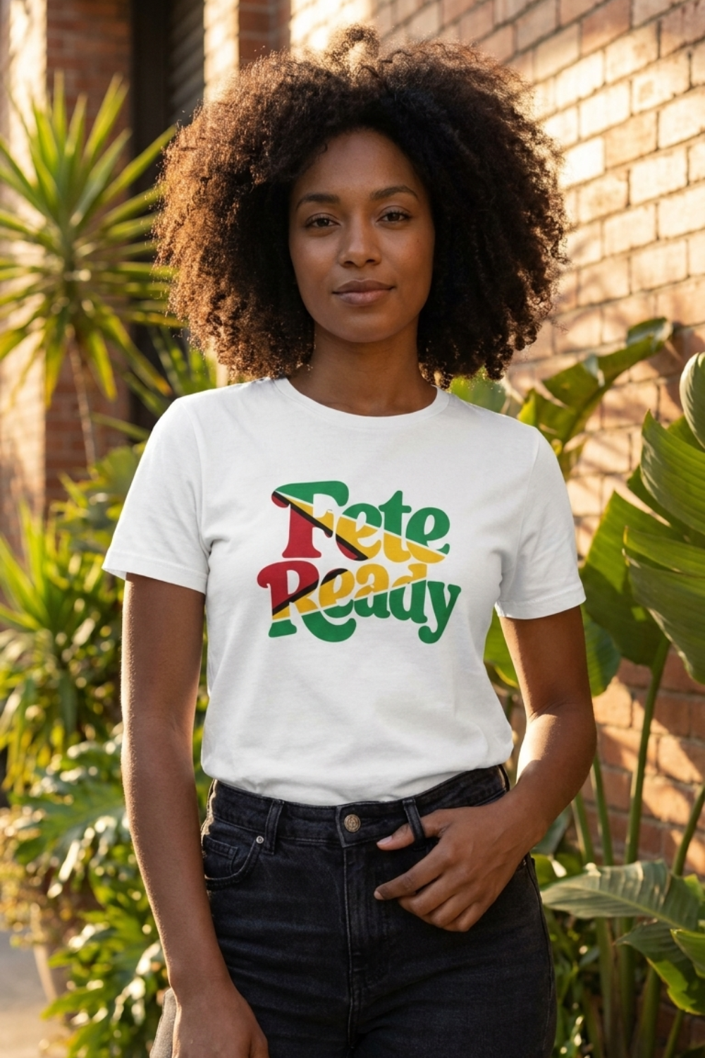 A woman with voluminous curly hair wearing a white "Fete Ready" t-shirt with the Guyana flag colored retro lettering. She is wearing black jeans and standing outdoors against a brick wall with lush green plants.