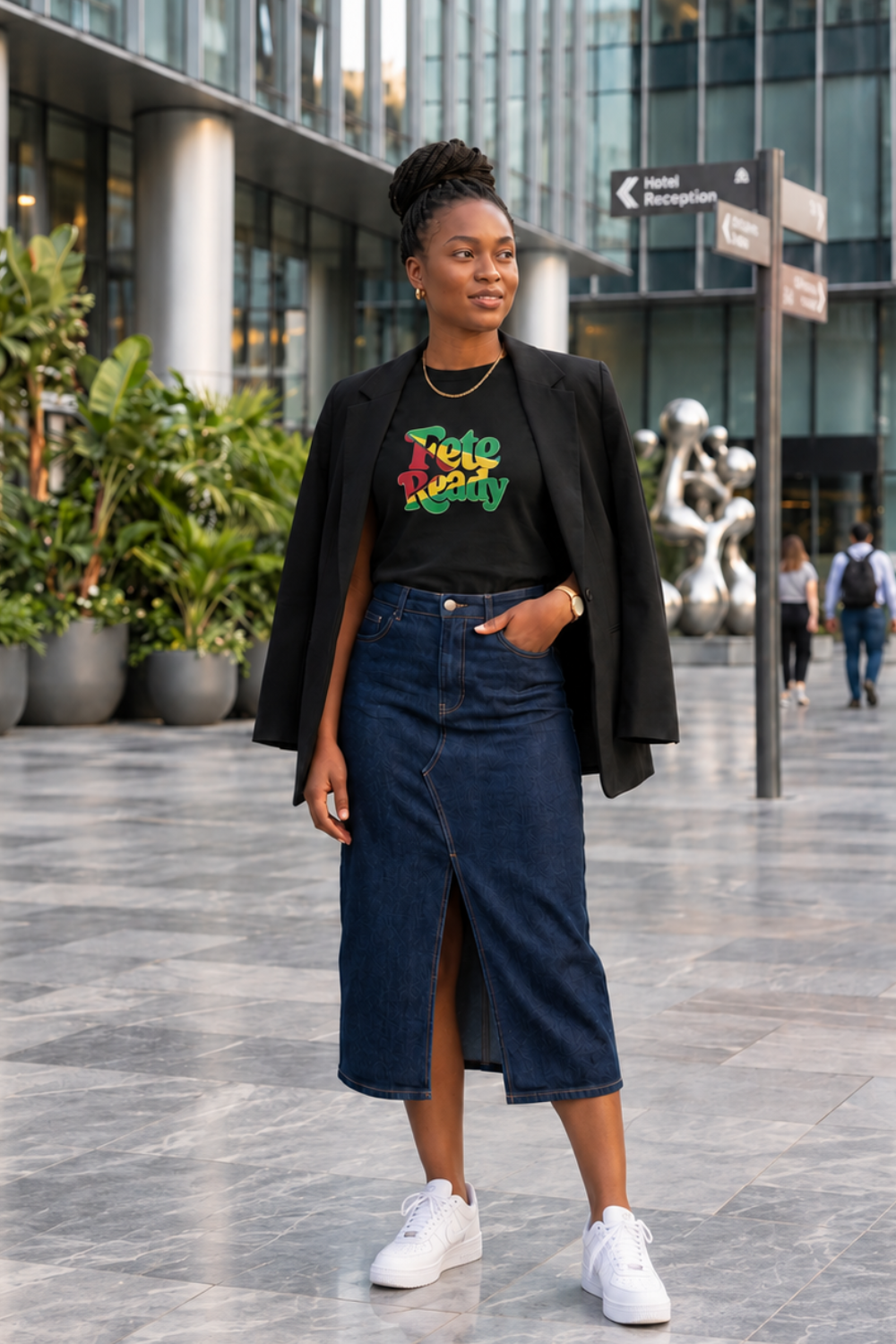 A woman stands outdoors in a modern plaza wearing a black graphic T-shirt with Fete Ready in the Guyanese flag colours a black blazer, a dark blue denim midi skirt with a front slit, and white sneakers.
