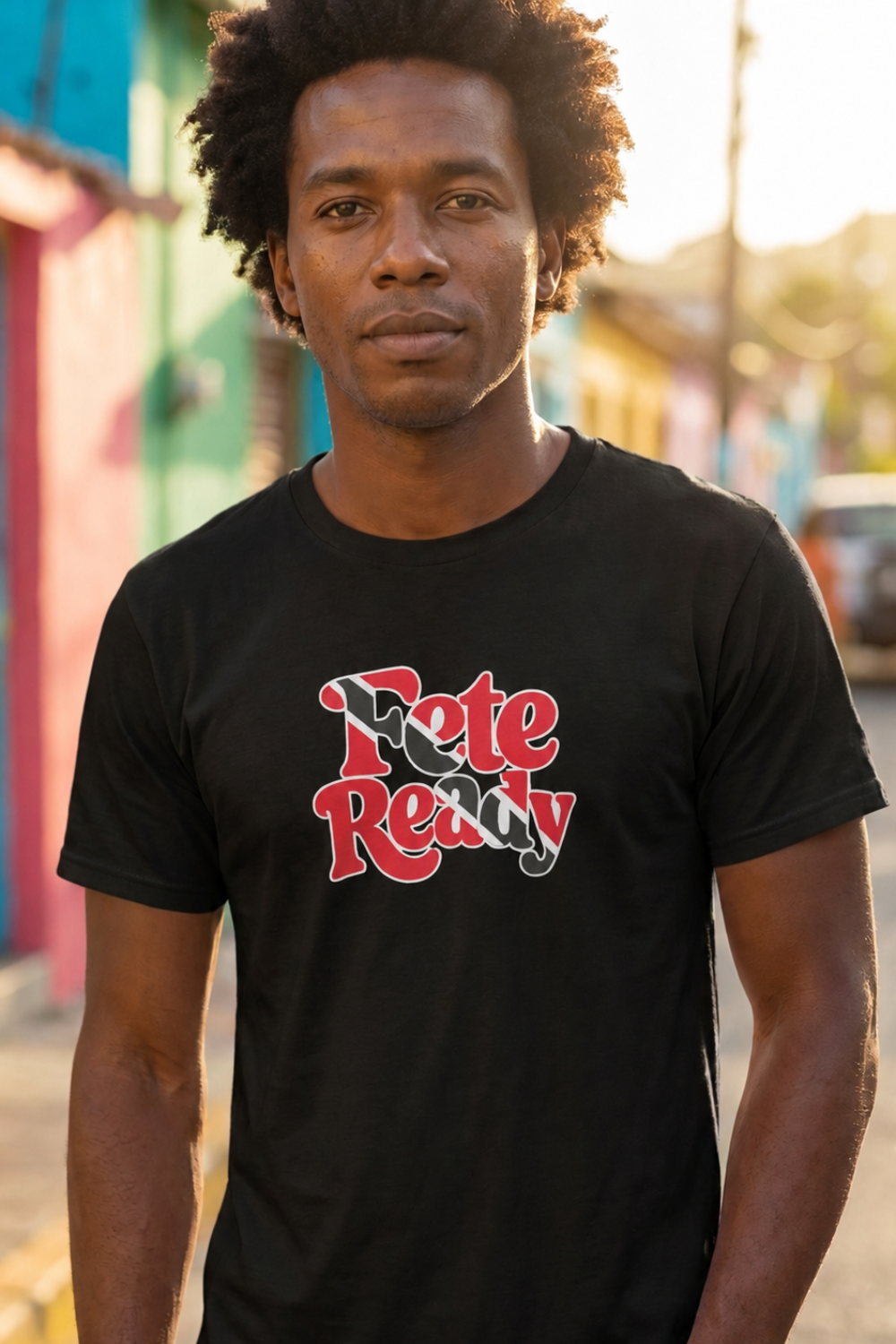 A man wearing a black crewneck t-shirt with a red, black and white (the Trinidad & Tobago flag colours) graphic text print that reads 'Fete Ready'. The background is a softly blurred outdoor street scene with colorful buildings.