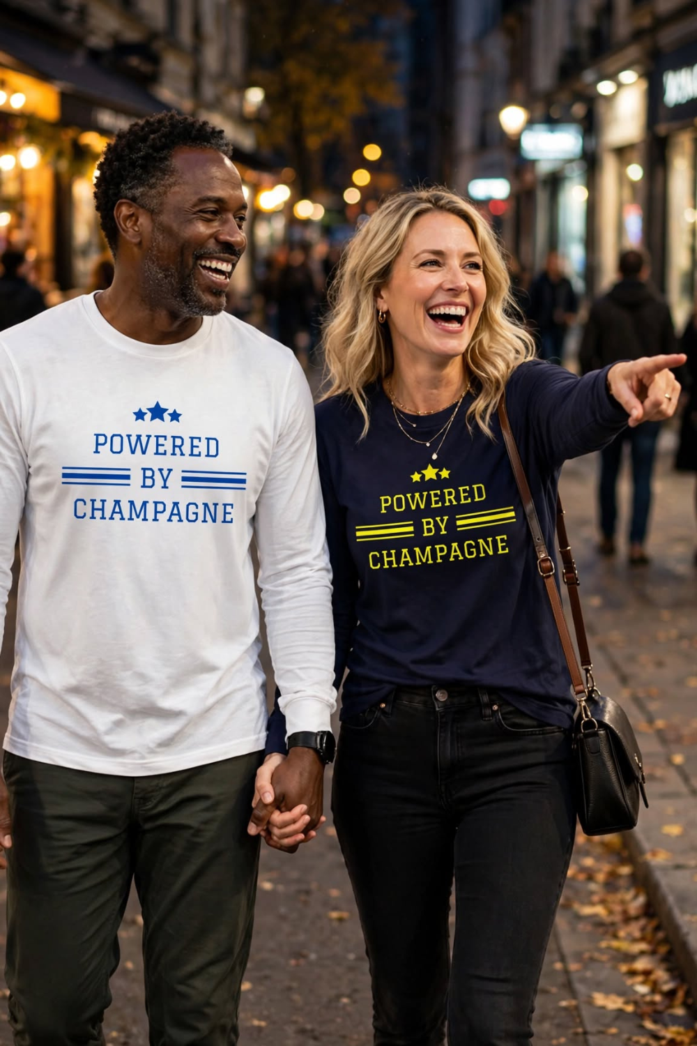 Interracial couple holding hands and laughing on fall city street wearing Powered by Champagne long sleeve tees - white unisex long sleeve with blue text and navy unisex long sleeve with yellow text - champagne lovers duo set