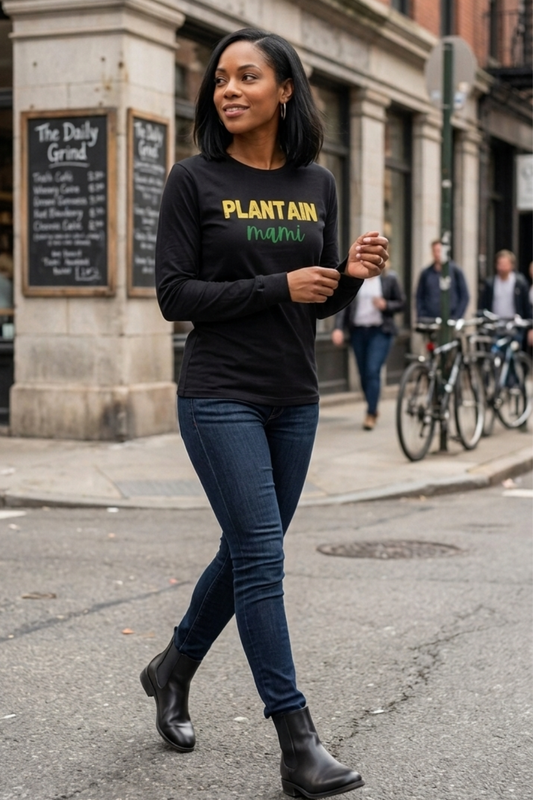 Black woman walking on a city street wearing a black long-sleeve Plantain Mami shirt with yellow and green lettering, dark jeans, and black ankle boots.
