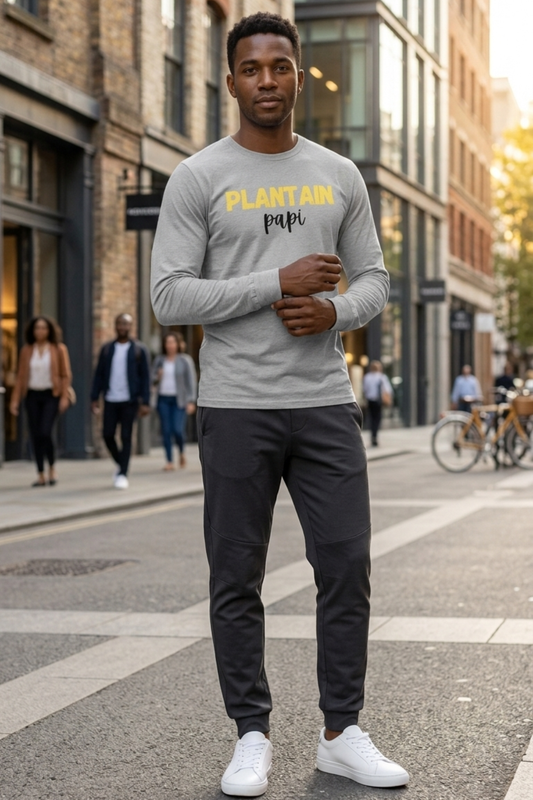Black man wearing a heather grey Plantain Papi long-sleeve shirt with yellow and black lettering, styled with dark tapered track pants and white sneakers on a city street.