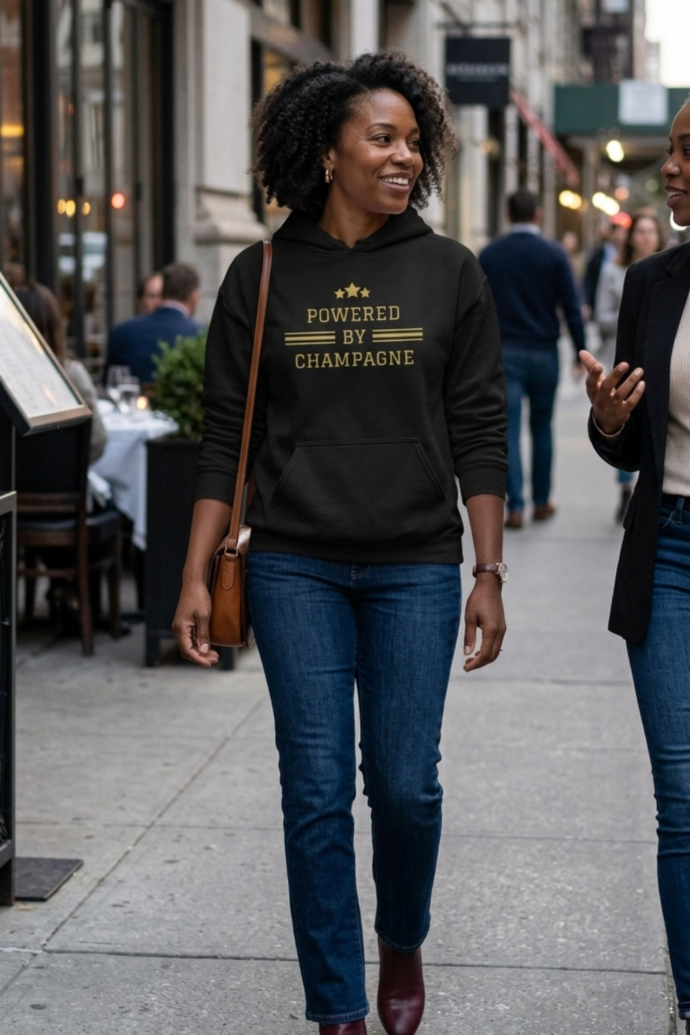 A woman walking on a city sidewalk wearing a black hoodie with gold text that reads 'POWERED BY CHAMPAGNE' and blue jeans. Another person is partially visible at the right edge of the frame.