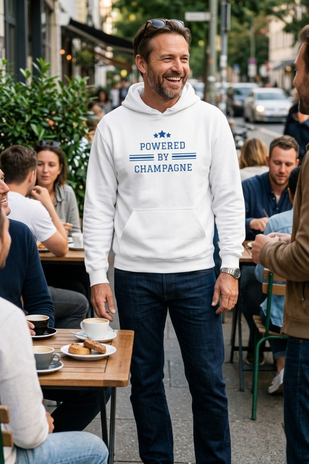 A man standing at an outdoor cafe wearing a white hoodie with blue text that reads 'POWERED BY CHAMPAGNE' and dark blue jeans, surrounded by seated customers and tables with coffee.