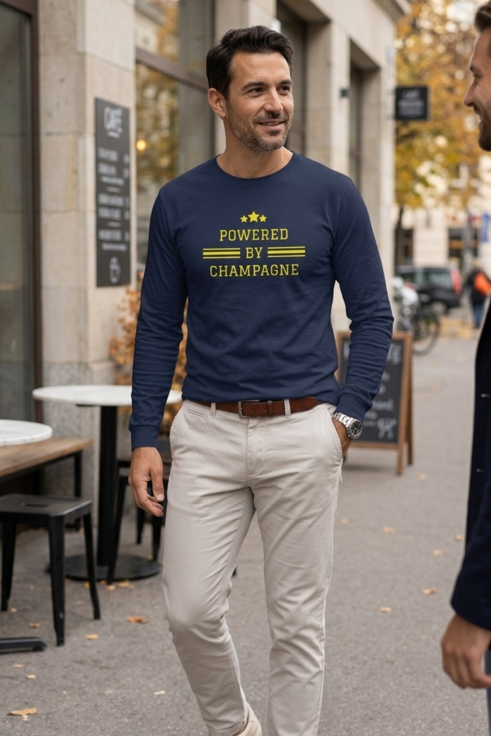 Man wearing a navy blue Powered by Champagne long-sleeve wine t-shirt by Gourmandee Tees, featuring gold varsity-style lettering, three stars and horizontal stripes. Styled with beige trousers on a city street outside a café.