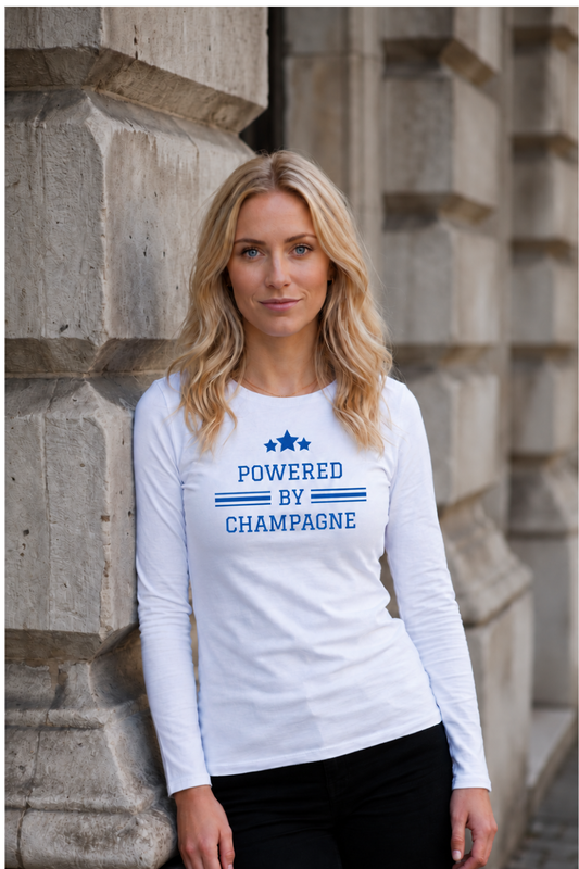 Blonde woman wearing a white Powered by Champagne long-sleeve wine t-shirt by Gourmandee Tees, featuring blue varsity-style lettering and three stars. Styled with black trousers against a stone building.