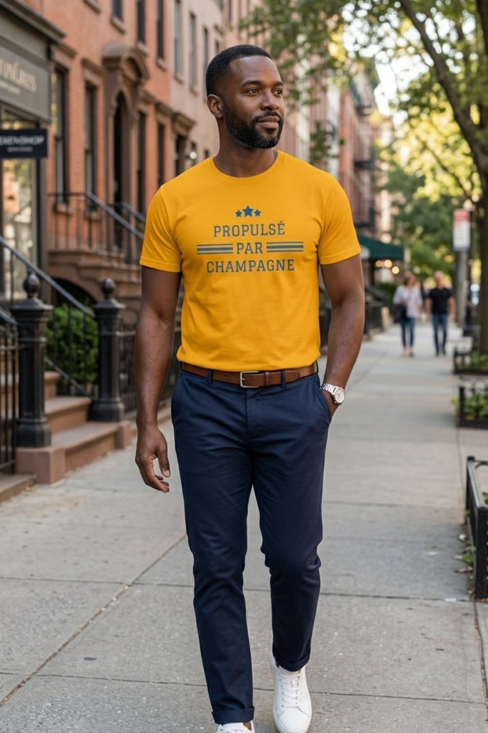 Premium golden-yellow unisex short-sleeve champagne t-shirt featuring “Propulsé par Champagne” in contrasting blue lettering, with three blue stars and horizontal stripes. Black male model styles the crewneck French wine lifestyle tee with navy trousers and white sneakers on a tree-lined city street.