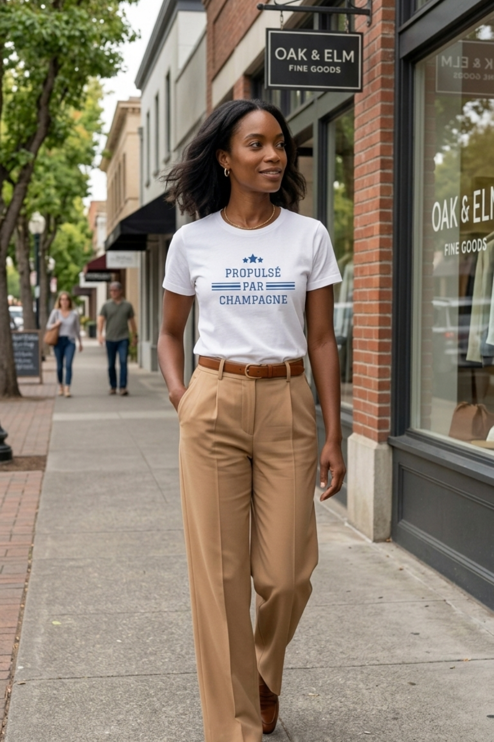 Premium white unisex short-sleeve champagne t-shirt featuring the French phrase “Propulsé par Champagne” in blue lettering, with three blue stars and horizontal stripes. Black female model wears the crewneck wine lifestyle tee tucked into high-waisted camel trousers with a brown leather belt while walking past city boutiques.