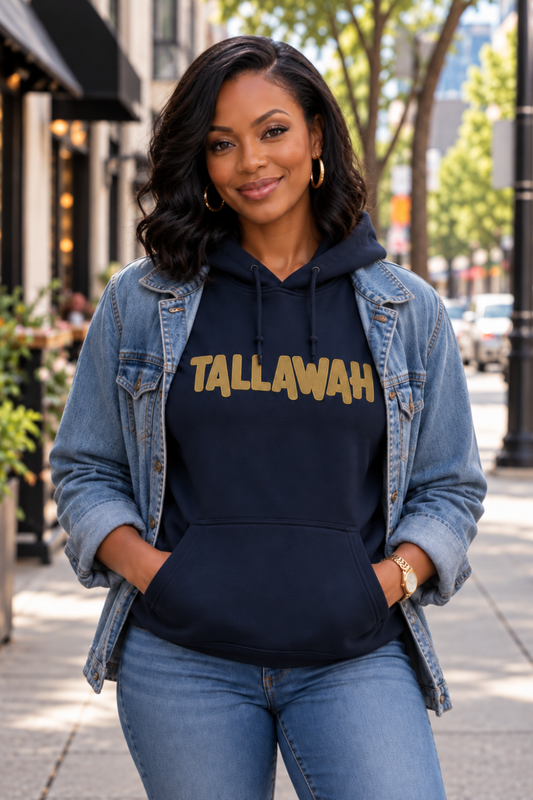 black model wearing a navy hoodie with front pockets and the word "TALLAWAH" embroidered on old gold on the front. This is Jamaican clothing. The model is wearing a blue jean jacket that is open over the hoodie and blue jeans. She standing on the sidewalk of a city street. Clothing designed to Rep Your Roots - Caribbean Apparel.