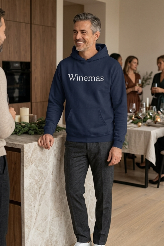Man wearing a navy blue pullover wine hoodie with “Winemas” printed across the chest in large white serif lettering. The hoodie features long sleeves, ribbed cuffs, an adjustable hood and a front kangaroo pocket. Styled with charcoal trousers at an indoor Christmas dinner party.