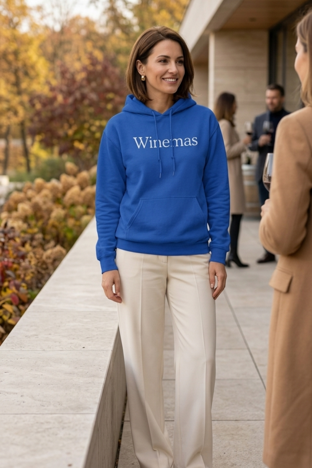 Woman wearing a royal blue pullover wine lover hoodie with “Winemas” printed in white serif lettering across the chest. Featuring a drawstring hood, long sleeves, ribbed cuffs and a front kangaroo pocket, the hoodie is styled with cream wide-leg trousers at an outdoor autumn gathering.